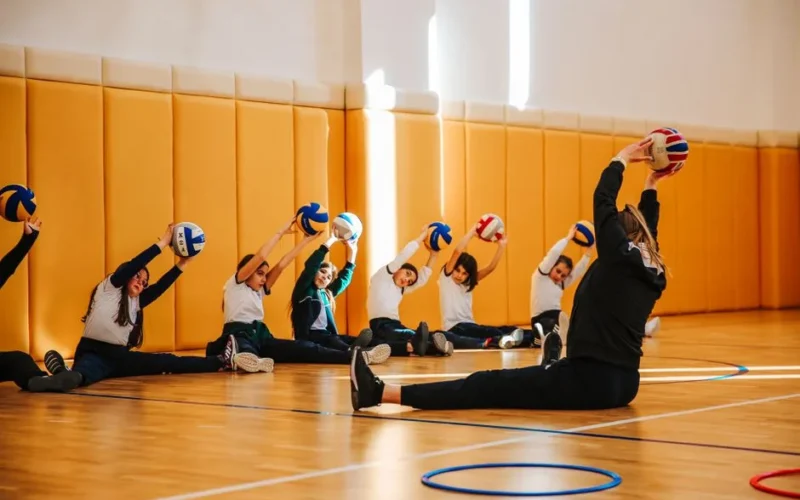 Group of children practicing volleyball stretches in a spacious indoor gym with a coach.