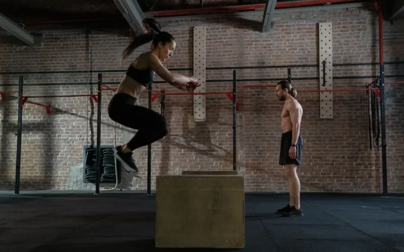 Female athlete performing box jump in gym watched by coach. Intense fitness training indoors.