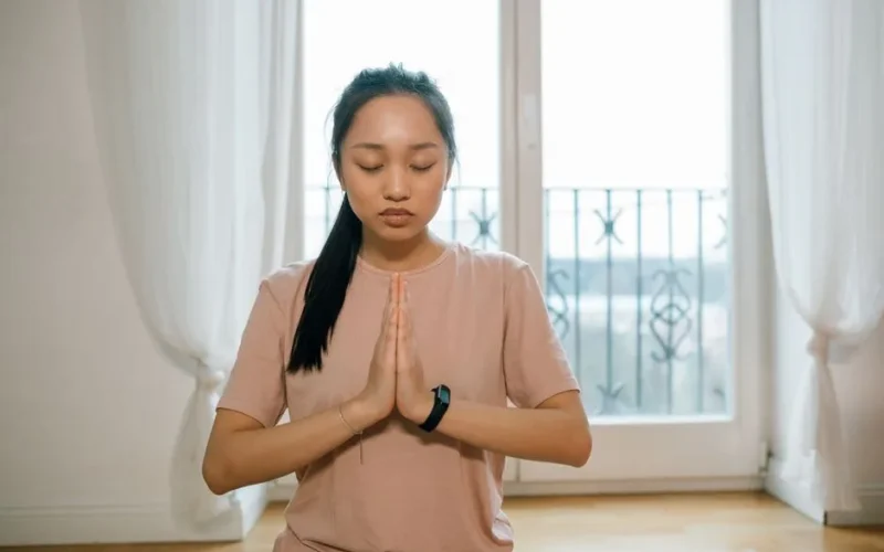 Asian woman practicing meditation indoors, hands together, promoting peace and healthy lifestyle.