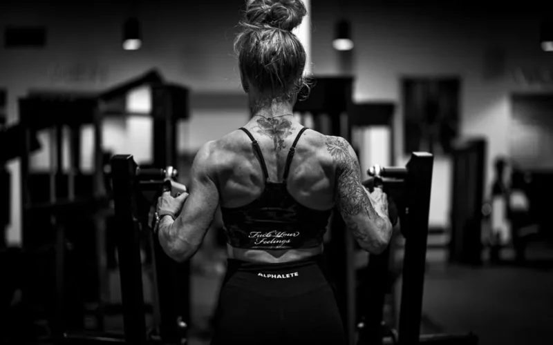 Back view of a tattooed woman lifting weights in a gym, showcasing strength and physique.