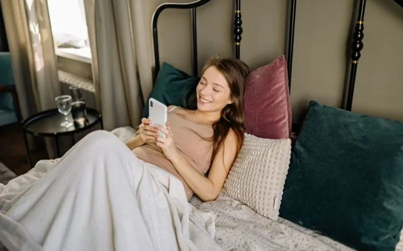 Smiling woman relaxes with smartphone in a cozy, well-lit bedroom setting.