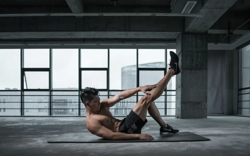 A shirtless man performs an ab workout on a yoga mat in an urban indoor gym setting.
