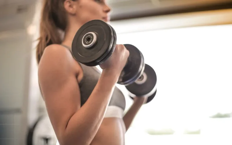 Focused woman lifting dumbbells in a bright gym, emphasizing strength and fitness.