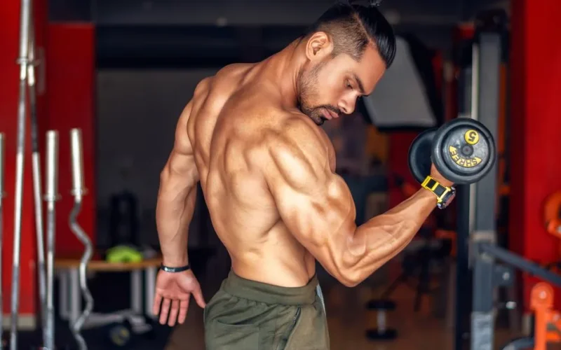 A fit, shirtless man showcasing biceps while lifting a dumbbell in a gym setting.
