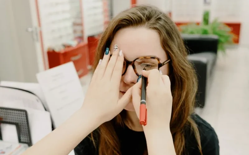 Anonymous female doctor covering eye of female client while choosing proper lenses during appointment in optical salon on blurred background
