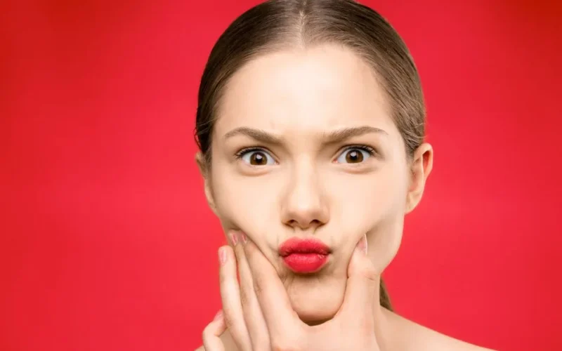 A young woman squeezes her cheeks, making a funny face against a vivid red background.