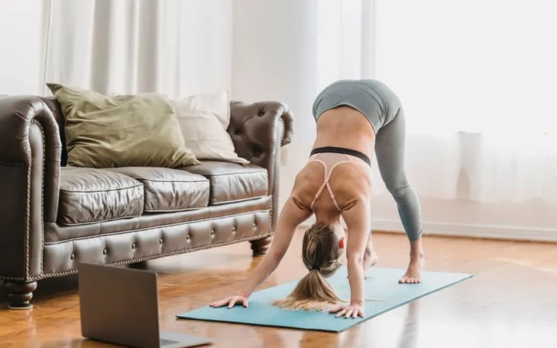 Full length of faceless lady wearing leggings and sportive bra stretching back on yoga mat near opened netbook and leather couch with pillows in light living room near curtains