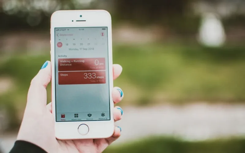 A person holds a smartphone showing a fitness app with step count and distance tracking outdoors.