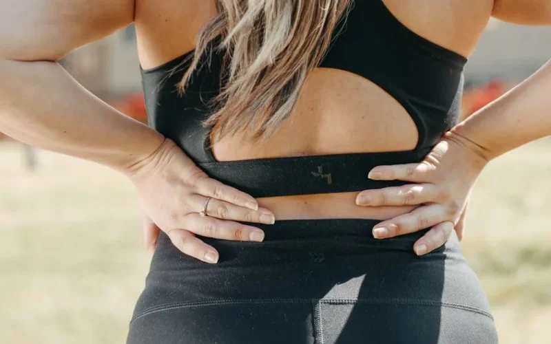 A woman in activewear holds her lower back, indicating muscle strain, outdoors.