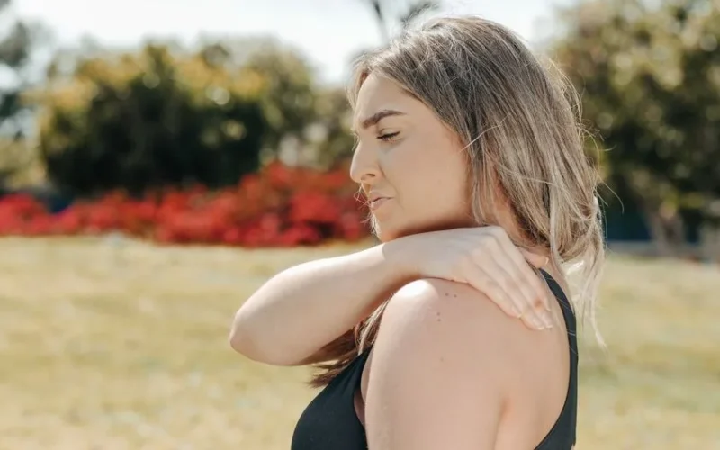 Woman in black tank top touching shoulder outdoors. Concept of shoulder pain and relaxation.