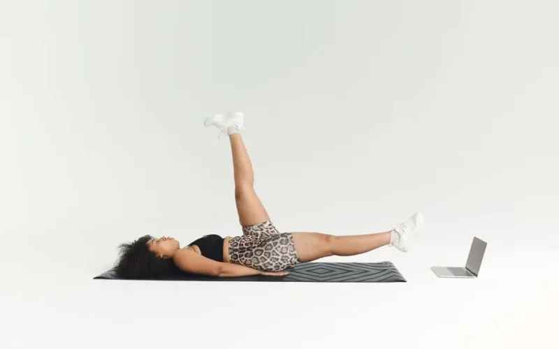African American woman exercising indoors on a mat with a laptop for an online workout session.