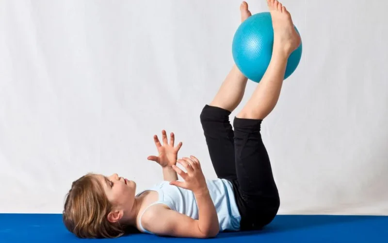 Young girl practicing balance exercises with a blue yoga ball indoors on a mat.