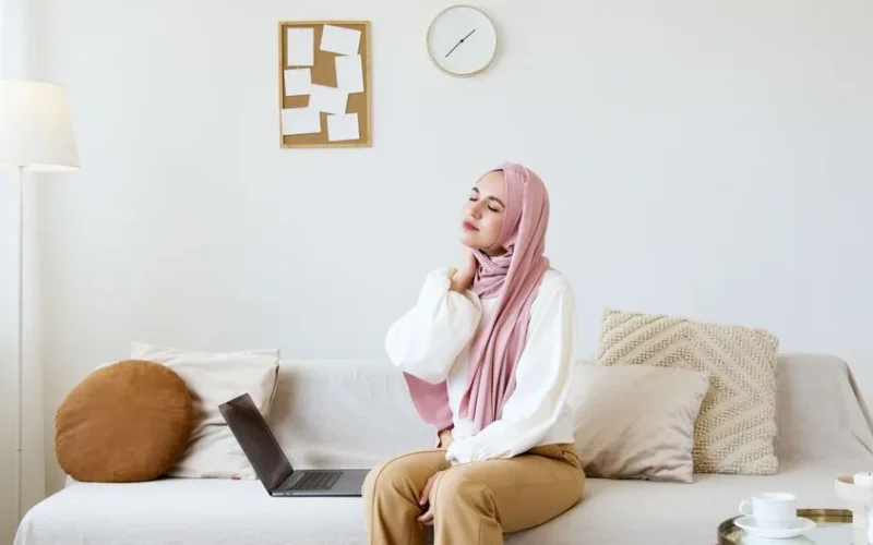 Muslim woman in hijab sitting on sofa, relaxing with eyes closed and hand on neck. Modern living room setting with laptop.