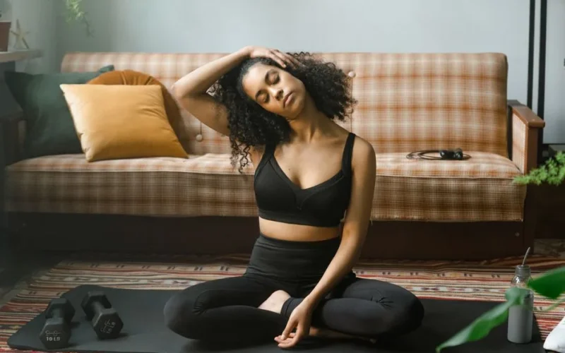 Woman in activewear performing a neck stretch on a yoga mat indoors, promoting a healthy lifestyle.