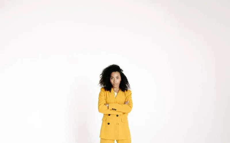 Portrait of a confident woman in a yellow suit with curly hair, arms crossed, posing in a studio setup.