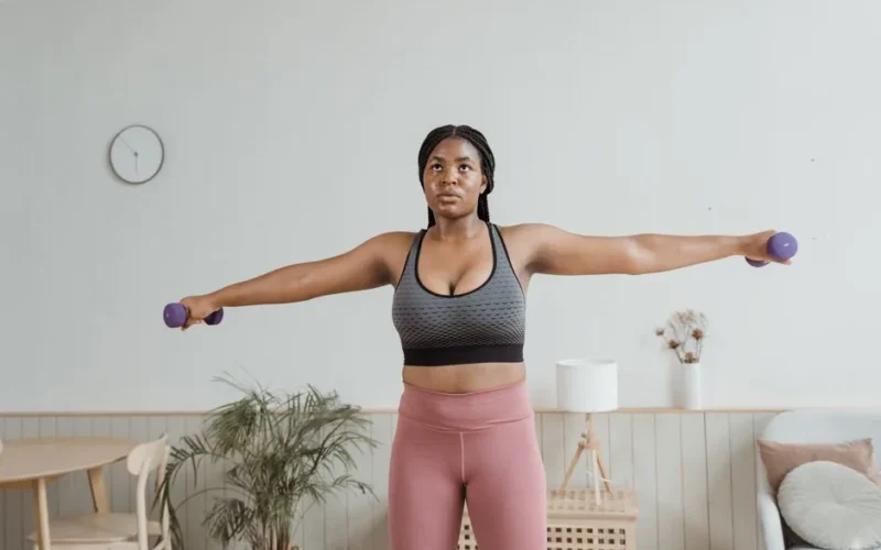 Black woman in activewear performing arm exercises indoors with dumbbells.