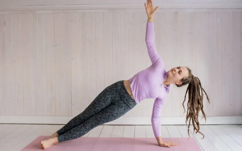 Woman in a side plank yoga pose on a pink mat in a bright indoor space.