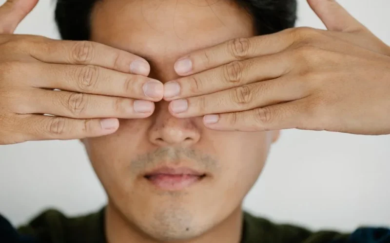 Close-up portrait of a man covering eyes with both hands against a plain background.