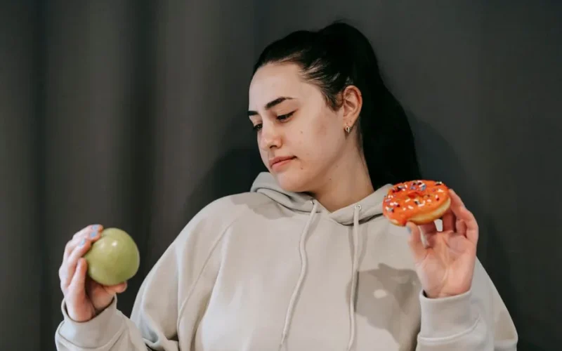 Pensive female wearing casual hoodie deciding between healthy green apple and sweet doughnut in studio