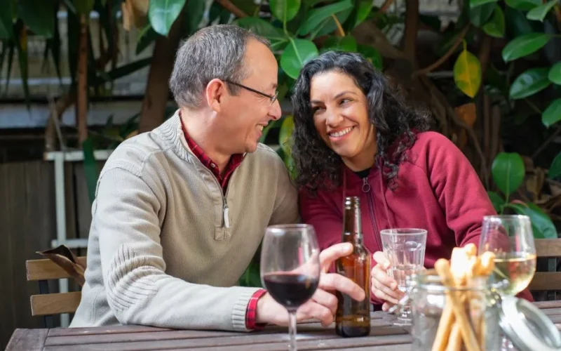 A cheerful couple sitting at a table, sharing smiles over wine and beer in an outdoor backyard.