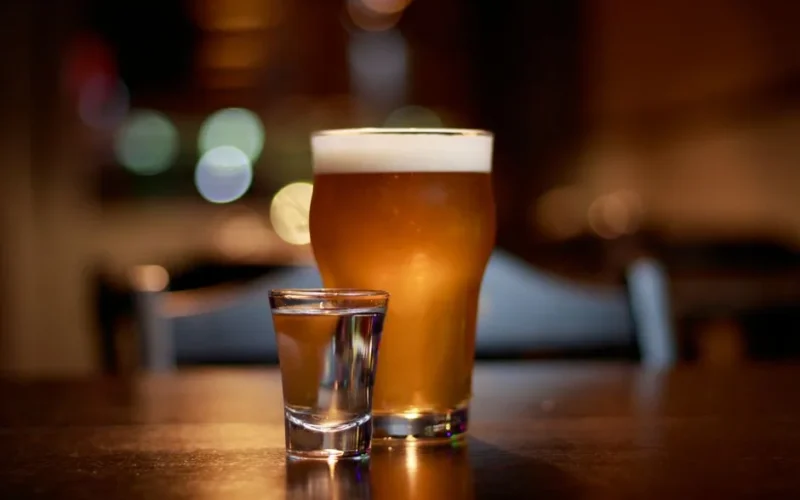 A close-up of a beer glass and vodka shot on a pub table with bokeh lighting.
