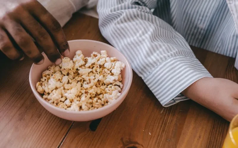 Close-up of hands grabbing popcorn from a ceramic bowl on a wooden table.