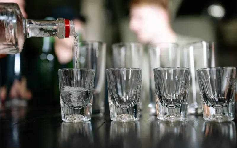 Close-up of a bottle pouring clear alcohol into shot glasses, with people in the background.