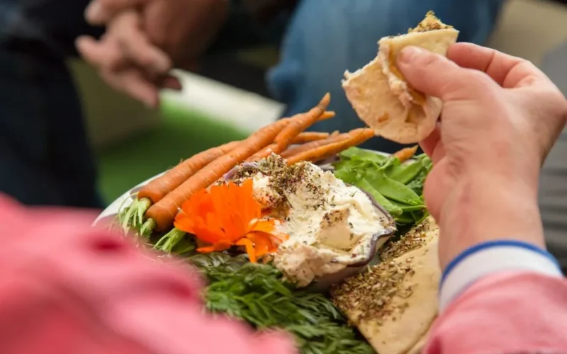 Close-up of hands enjoying hummus with fresh vegetables and pita bread, showcasing healthy eating.