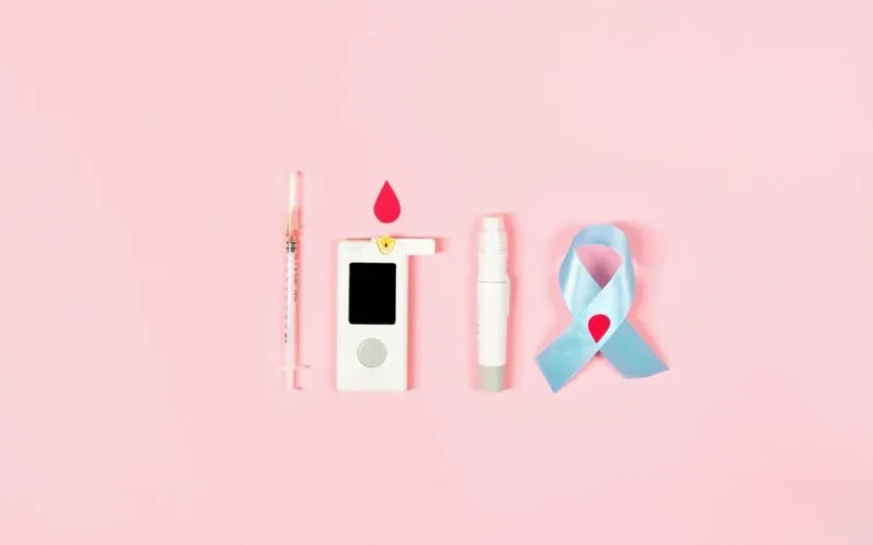 Glucometer, syringe, lancet pen, and ribbon symbolizing diabetes awareness on pink background.