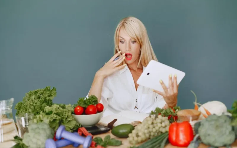 A surprised woman looking at a tablet surrounded by fresh vegetables and fruits, promoting healthy eating.