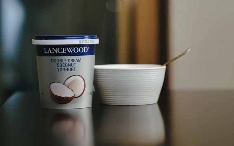 Lancewood double cream coconut yoghurt with a spoon and bowl on a reflective surface.