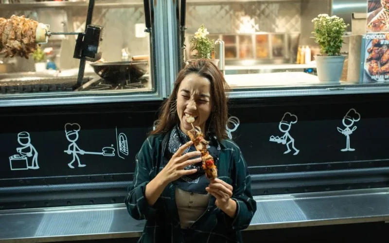 Smiling woman biting into a delicious kebab at a vibrant street food truck.