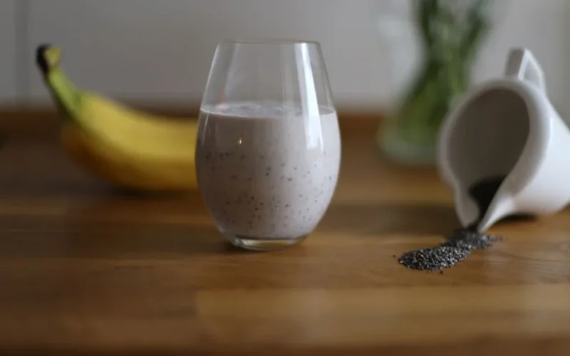 Glass of chia seed smoothie with banana and spilled seeds on wooden table.