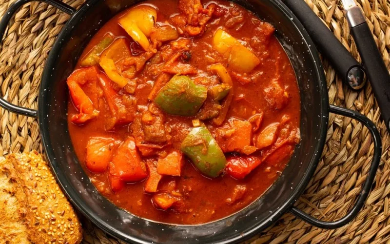 Delicious bell pepper stew served in a black pot with sesame seed bread on a woven placemat.