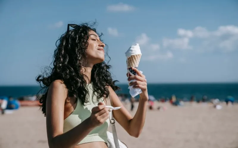 A woman with curly hair enjoys a vanilla ice cream cone at the beach on a sunny summer day.