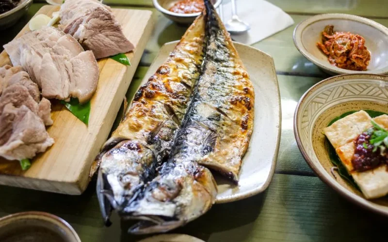A top view of a platter with sliced pork, grilled mackerel, kimchi, and other Korean dishes on a wooden table.