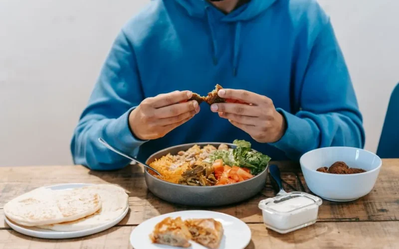 Crop unrecognizable male tasting fried meat from dish with fresh vegetables at wooden table with plates filled with food