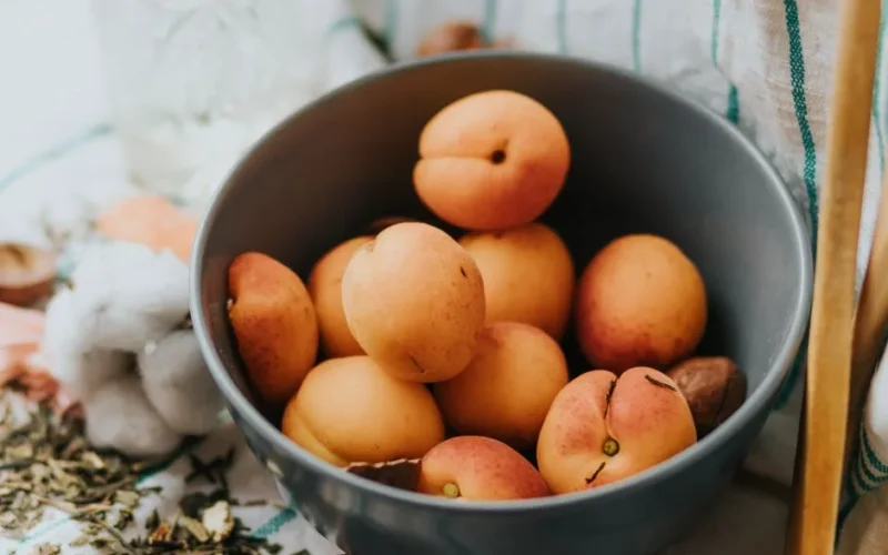 A close-up shot of fresh apricots in a gray bowl on a kitchen table, surrounded by herbs.