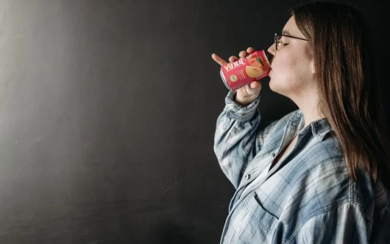 A woman in a casual shirt drinks from a canned beverage against a dark background.