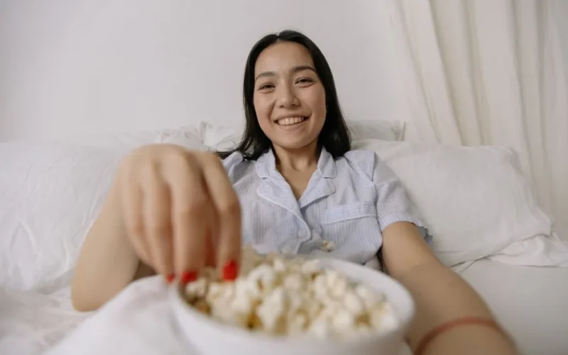 Asian woman in pajamas smiling while eating popcorn in a cozy bedroom setting.
