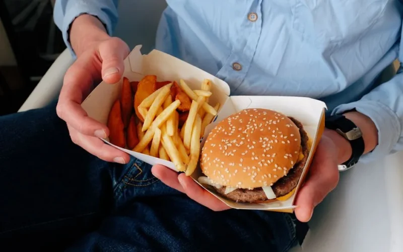 Unrecognizable male with wristwatch in casual clothes sitting on chair and holding box of burger and french fries in hands