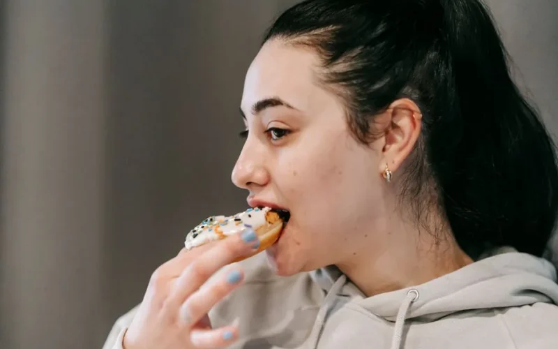 Crop young brunette in comfy hoodie eating fresh appetizing doughnut covered with vanilla icing and sprinkles in light room