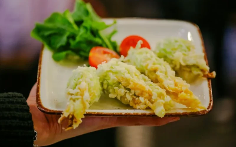 Crispy tempura zucchini blossoms served with fresh tomato and leafy greens on a square plate.