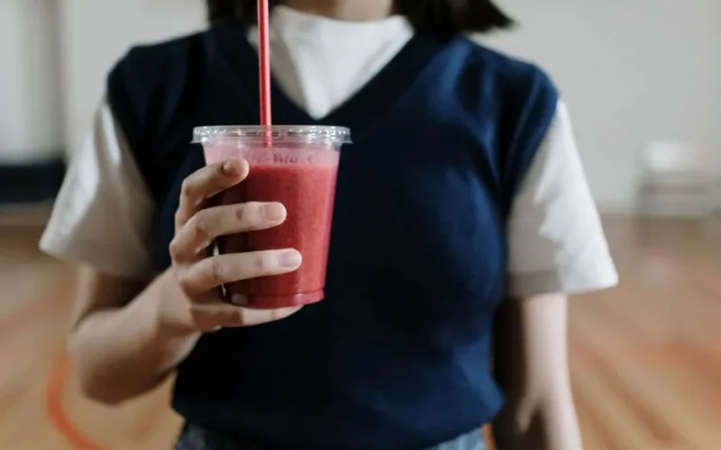 Close-up of a woman holding a red fruit smoothie in a plastic cup indoors.