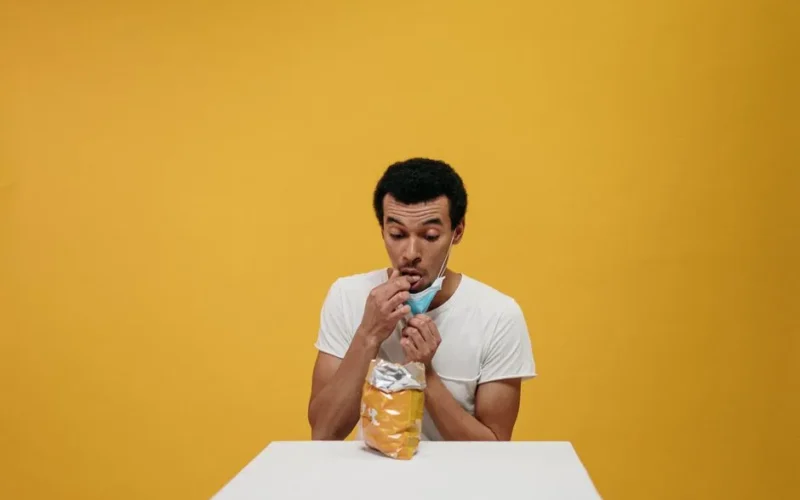 Man humorously eating chips while wearing a mask in a studio setting.