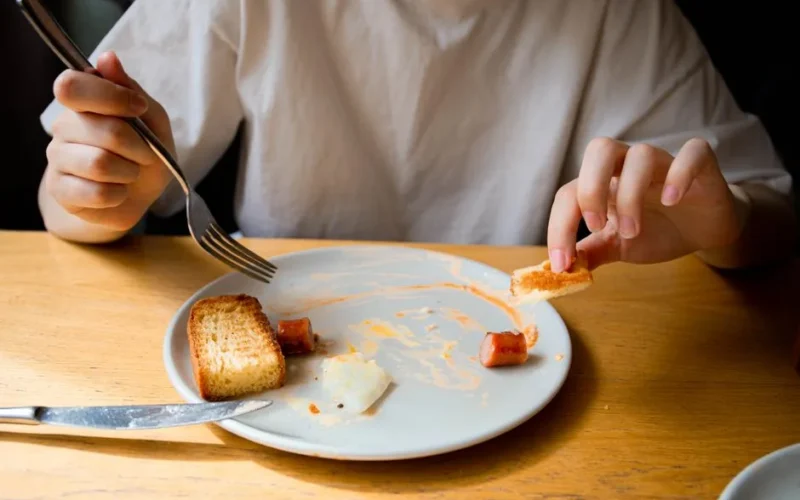A person enjoying a casual meal of toasted bread and sausage on a white plate at a wooden table.