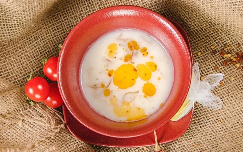 Top view of creamy red lentil soup with tomatoes and olive oil in a red ceramic bowl on a burlap background.