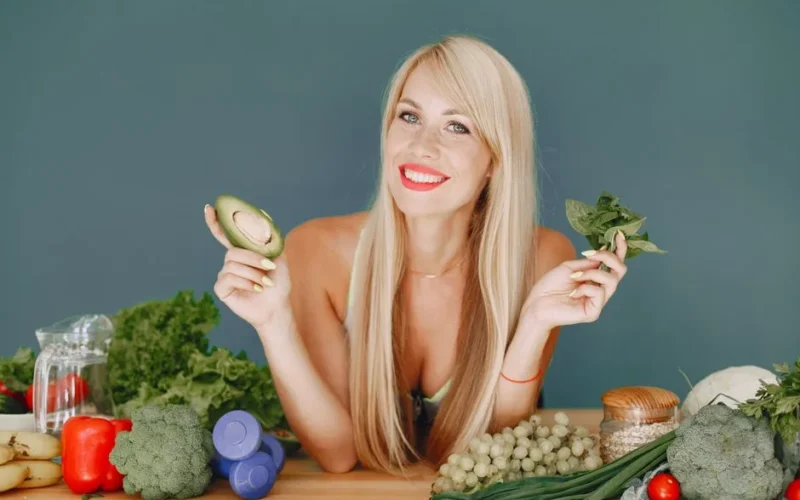 Blonde woman promotes healthy eating with fresh vegetables and fruits on display.