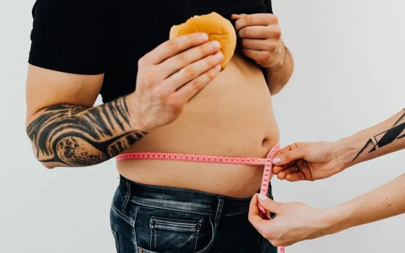 A man with tattoos holding a burger as another person measures his waistline with a tape measure.