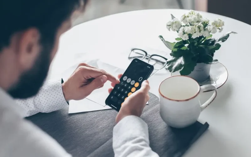 Adult man using smartphone calculator at table with flowers and coffee cup.